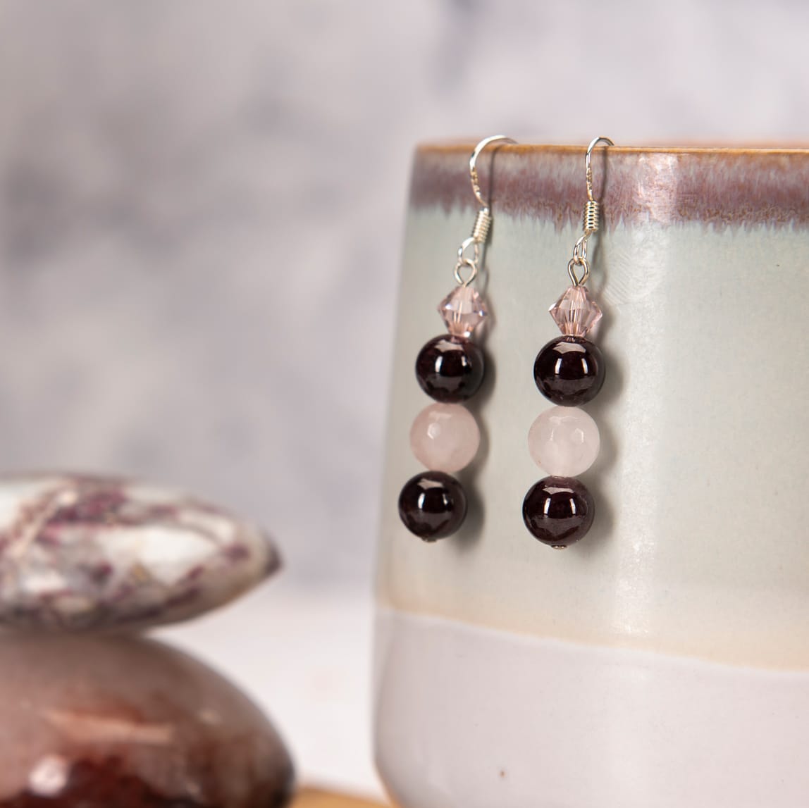 A pair of beaded dangle earrings with deep red, pale pink, and clear faceted crystal beads on silver fishhook wires, hanging from the rim of a ceramic cup.