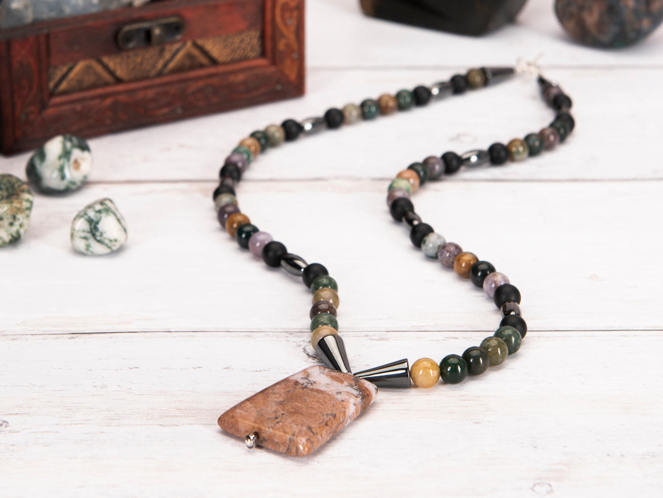 Necklace with multicolored beads and stone pendant on a wooden surface with rocks and a box in the background.