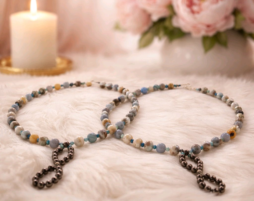 A pair of matching blue and silver necklaces on white fur with a candle and flowers in the background.