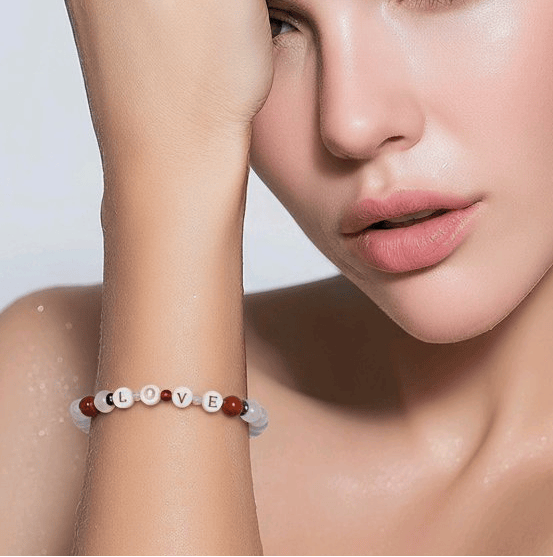 A close-up lifestyle shot of a model wearing a beaded bracelet on her wrist. The bracelet features white round beads with raised letter charms spelling 'LOVE' in gold letters, accented by small deep orange and pink beads.