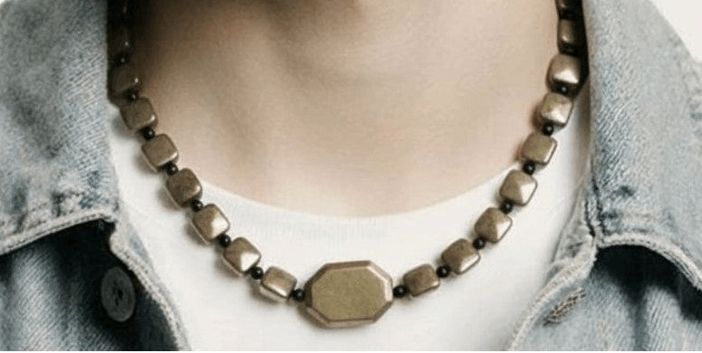 A close-up of a person wearing a chunky geometric bronze necklace with a central octagonal pendant, styled with a light blue denim shirt over a white t-shirt.