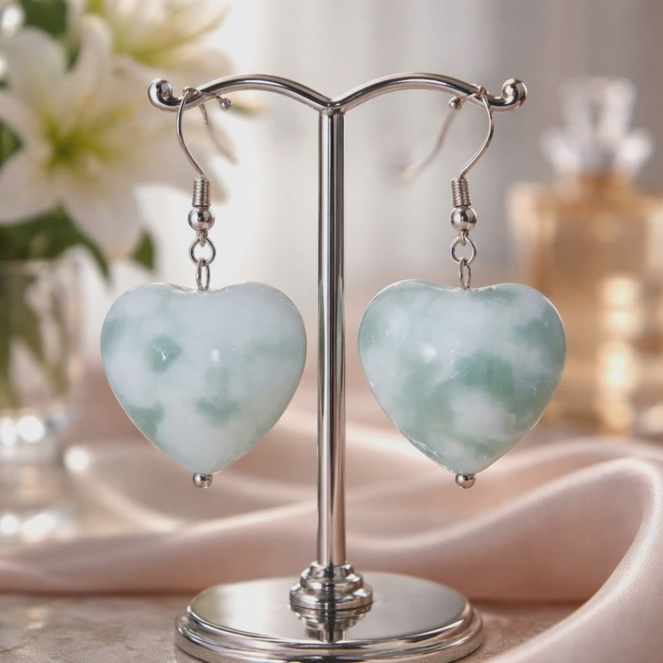 Video of green heart shaped earrings on a silver stand with flowers in the background.