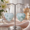 Video of green heart shaped earrings on a silver stand with flowers in the background.